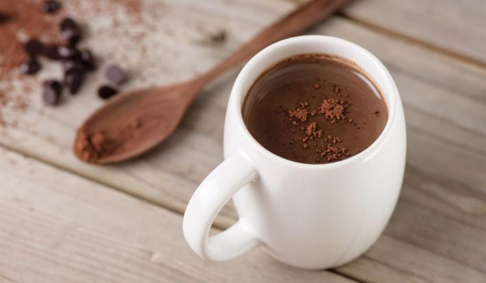Imagem de uma xícara branca com chocolate quente e ao lado uma colher de pau pequena com chocolate em pó.